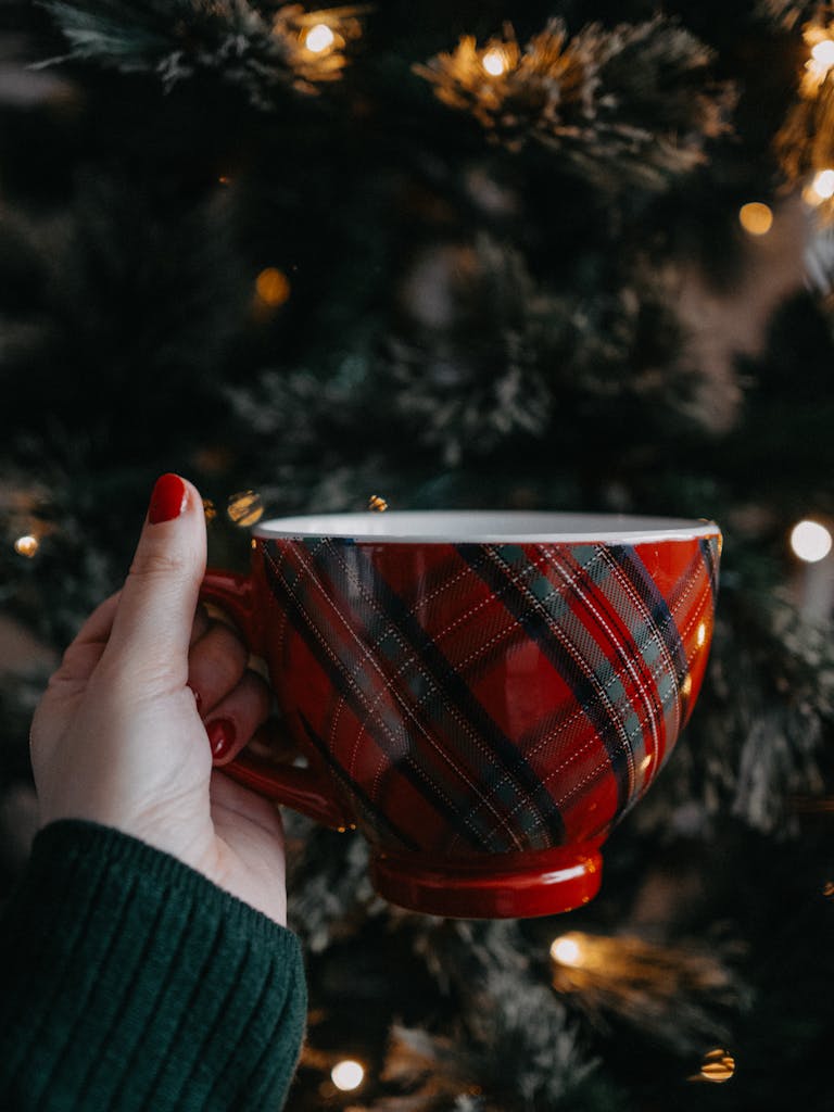 A hand holds a red plaid mug in front of a lit Christmas tree, embracing the festive warmth.