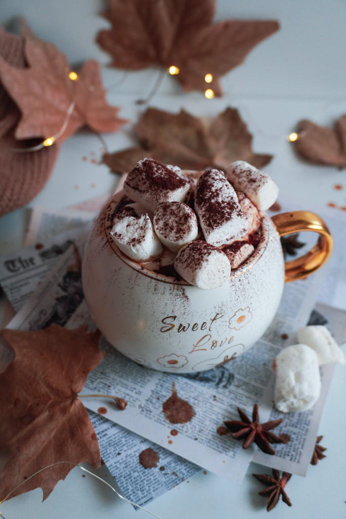 Warm mug of hot chocolate topped with marshmallows, surrounded by autumn leaves and cozy decor.